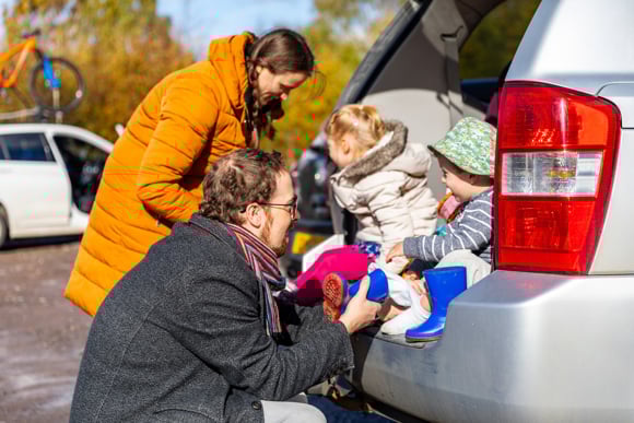 Happy family getting ready for a day out - Mother and father helping daughter and son sitting on car boot to wear boots