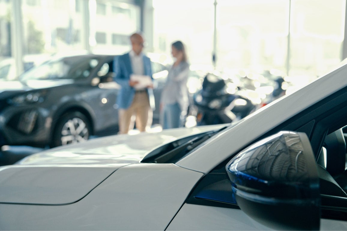 Close-Up of Brand New White Car with Female Customer and Male Salesperson Discussing in the Background at Modern Showroom