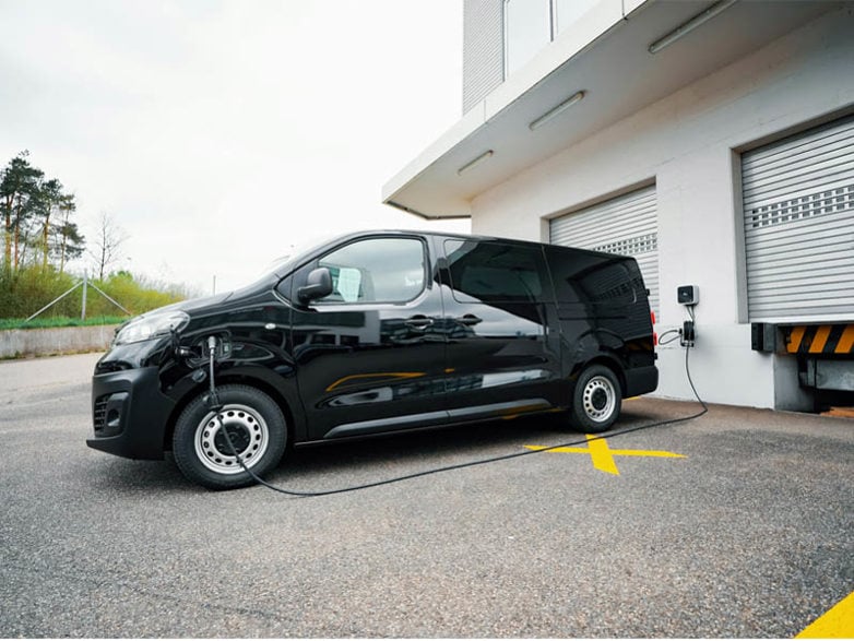 A black electric van plugged into charge at an industrial complex