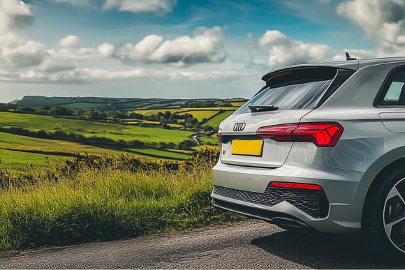 Rear view of a Audi hatchback parked on a rural road with rolling green hills and a partly cloudy sky in the background