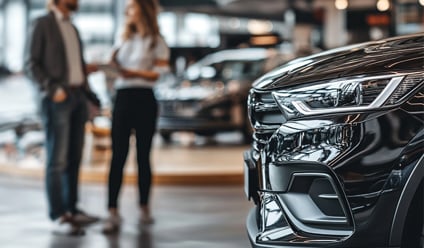 Close-up of a sleek black car in a modern dealership showroom, with two people in conversation in the blurred background