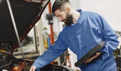 Mechanic in blue overalls looking an an engine bay with a tablet in his hand