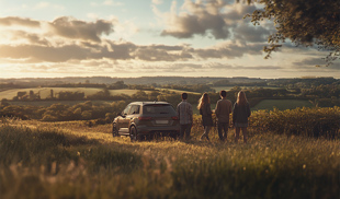 Group of young adults standing next to an SUV on a grassy hill at sunset, overlooking rolling countryside and fields