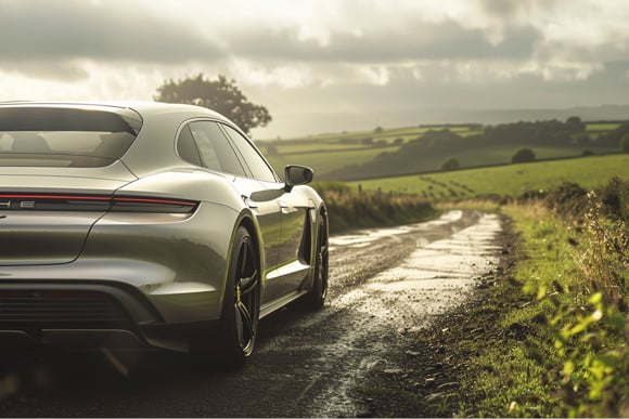 Rear view of a silver Porsche estate car on a rural countryside road with green fields and rolling hills in the background